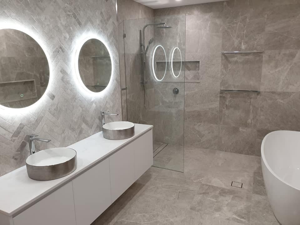 Modern bathroom with double vessel sinks, backlit circular mirrors, glass shower, and freestanding bathtub.